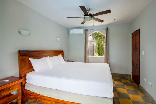 A simple, tidy bedroom with a large bed, wooden furniture, a ceiling fan, air conditioner, window, and door, in a white-painted room.