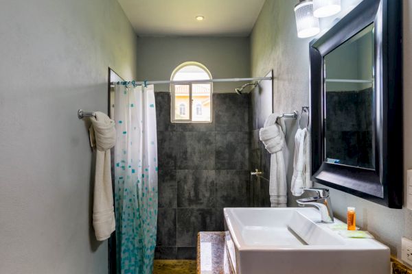 This image shows a bathroom with a rectangular sink, a large mirror, and three lights above it. There is a shower with a curtain and a window at the far end.