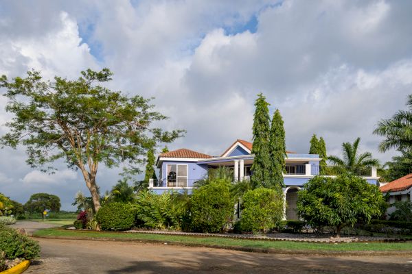 The image shows a two-story house with a red-tiled roof surrounded by lush greenery and trees under a partly cloudy sky.