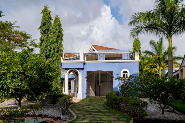 A blue two-story house with a gated entrance, surrounded by greenery, palm trees, and a paved pathway leading to the front door.