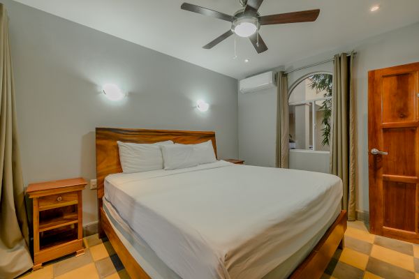 A bedroom with a large bed, two bedside tables, a ceiling fan, wall lights, a wooden door, curtains, and an air conditioning unit on the wall.