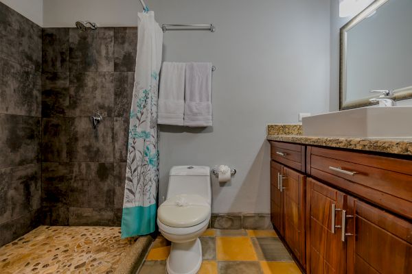 A bathroom with a tiled shower, white toilet, wooden vanity with granite countertop, sink, towel rack, and large mirror.
