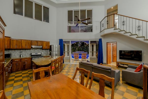 A spacious living area with a kitchen, dining table, sofa, TV, staircase, and balcony, featuring wooden furniture and checkered flooring.