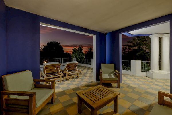 This image shows a patio with wooden furniture and cushioned seating, set against a blue wall, leading to an open view at sunset.