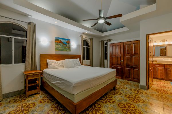 A bedroom with a large bed, ceiling fan, wooden furniture, patterned floor tiles, and an open door leading to a bathroom is shown.