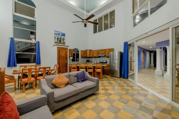 A spacious living area with high ceilings, a ceiling fan, a grey sofa, a dining table with chairs, and a kitchen with wooden cabinets and a stainless steel fridge.