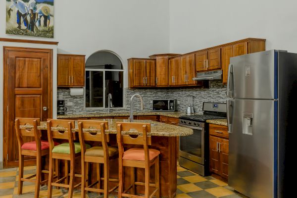 A modern kitchen with wooden cabinets, a stainless steel fridge, an oven, a microwave, an island with four chairs, and colorful cushions.