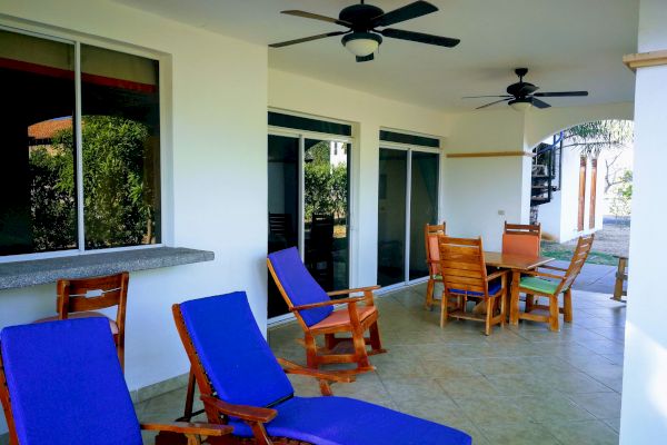 The image shows a patio with blue-cushioned lounge chairs, a wooden dining set, ceiling fans, and large sliding glass doors, suggesting an outdoor relaxation area.