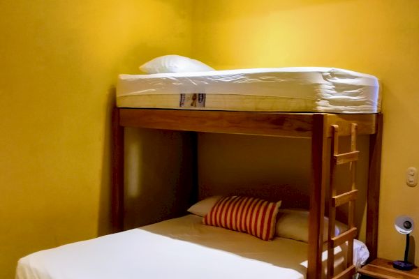 The image shows a cozy bedroom with yellow walls, featuring a bunk bed with a double bed below and a single bed above, and a ceiling fan.