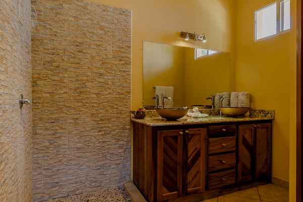 The image shows a bathroom with a walk-in shower, natural stone tiles, a wooden vanity with two sinks, and a large mirror above the vanity.
