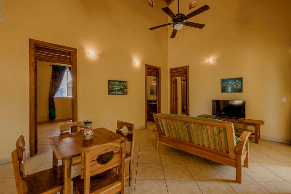 A cozy living room and dining area with wooden furniture, a flat-screen TV, ceiling fan, wall art, and doorways leading to other rooms.