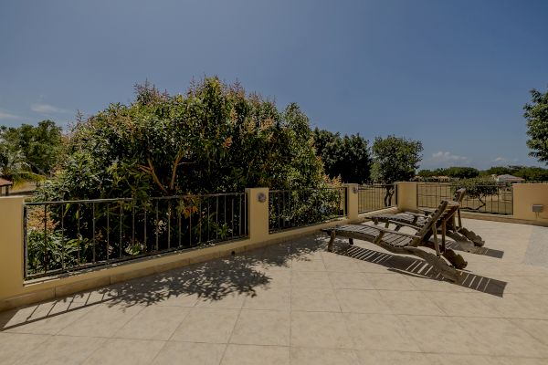 A sunlit patio with tiled flooring, lounge chairs, a garden with lush green trees and bushes, and a metal railing surrounding the area.