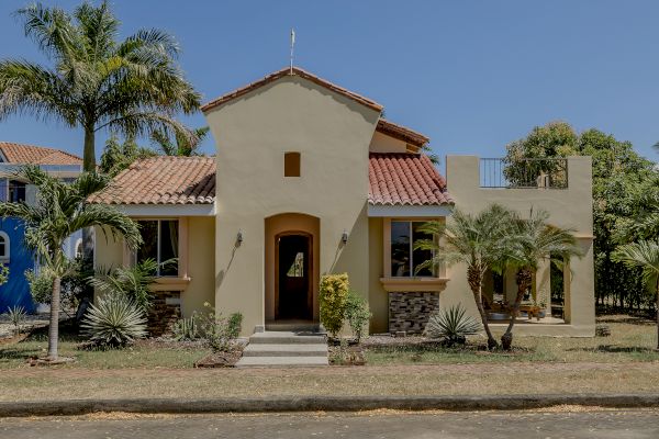 A beige stucco house with a red-tiled roof features a front entrance flanked by palm trees and desert plants, set against a clear blue sky.