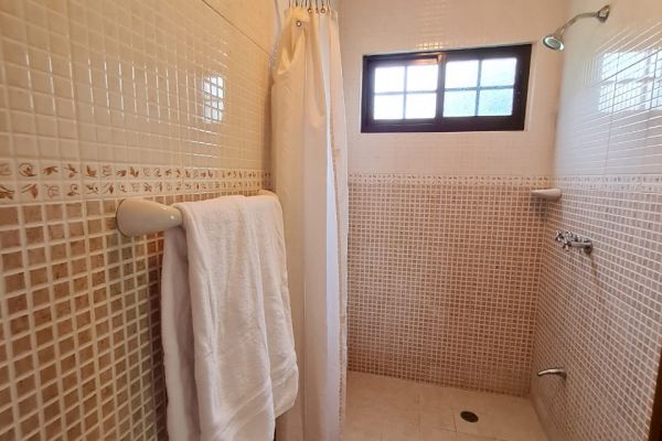 A bathroom with a walk-in shower, tiled walls, a white shower curtain, a window, a towel rack with white towels, and a dark floor mat.