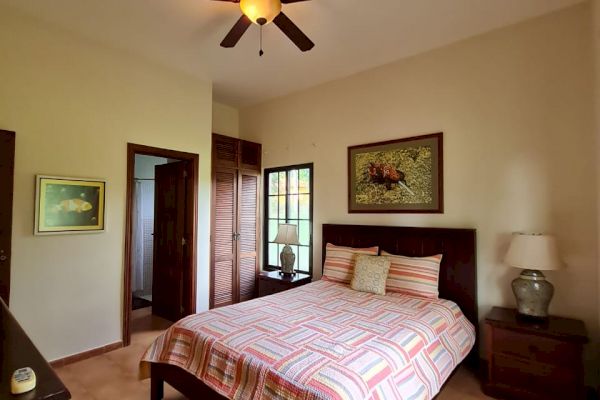A cozy bedroom features a bed with striped bedding, a ceiling fan, two wooden side tables with lamps, and framed wall art. Doorways lead to other rooms.