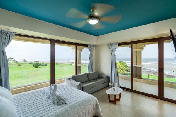 A modern, bright room with ocean views features a bed, a sofa, a coffee table, and large windows with blue curtains. The ceiling is painted blue.