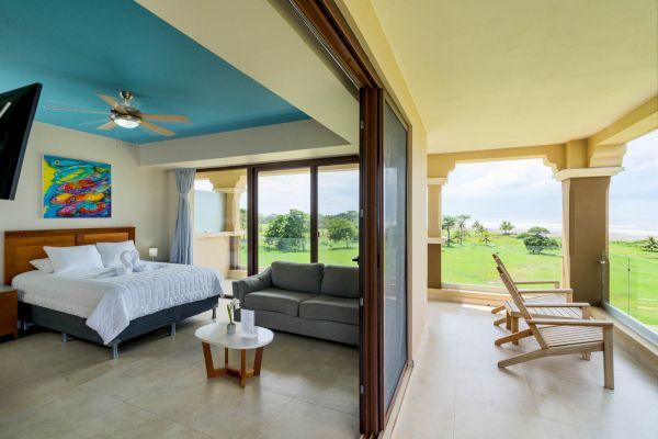A spacious bedroom with a bed, sofa, and colorful painting opens up to a balcony with lounge chairs and a scenic view of greenery and the ocean.