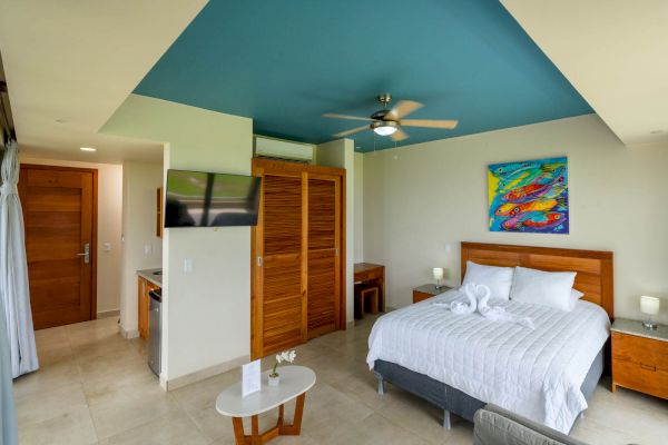 A modern bedroom with a blue ceiling, a large bed, a colorful painting above the headboard, a TV, and wooden furniture. White towels are on the bed.