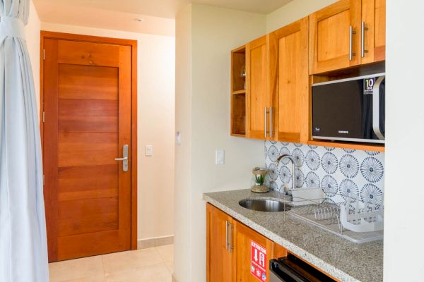 A compact kitchen with wooden cabinets, a small fridge, a microwave, a sink, and patterned backsplash tiles, leading to a wooden door.
