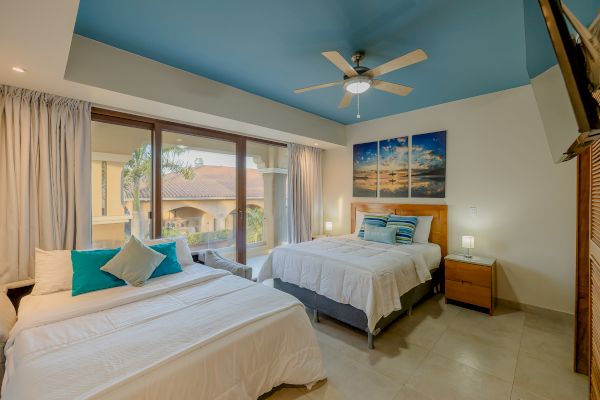 A bedroom with two beds, blue ceiling, wall art, a ceiling fan, bedside tables, lamps, and large windows with a view of a balcony and neighboring buildings.