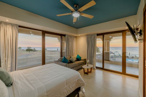 A bedroom with a double bed, sofa, ceiling fan, and wall-mounted TV, featuring large windows with an ocean view at sunset.