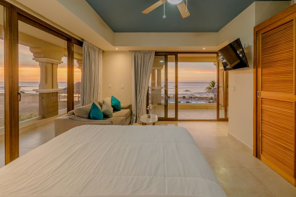 A cozy bedroom with a white bed, a small seating area, a wall-mounted TV, and large windows showcasing an ocean view at sunset.