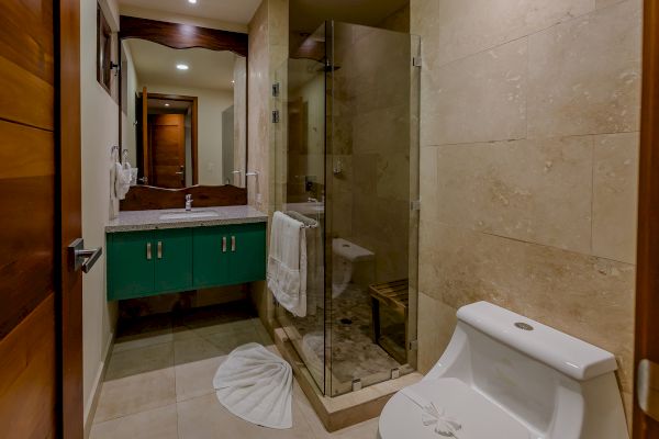 The image shows a bathroom with a green vanity, a sink, a glass-enclosed shower, and a white toilet. There's a towel and a folded mat on the floor.