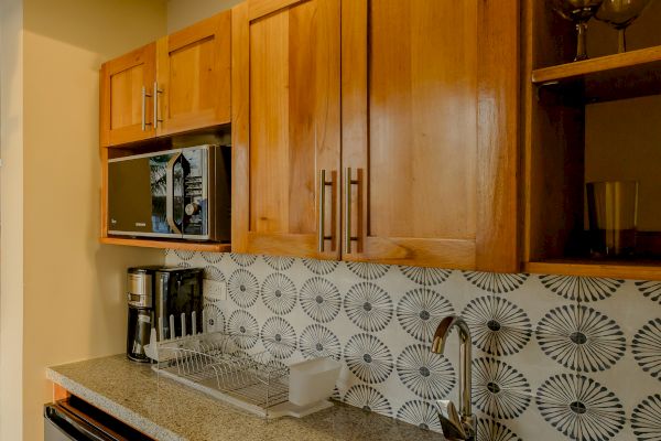 The image shows a small kitchen area with wooden cabinets, a granite countertop, a sink, a coffee maker, a microwave, and a tiled backsplash.
