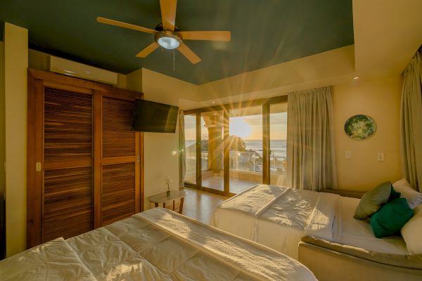 A cozy bedroom features two beds, a ceiling fan, a wall-mounted TV, and sliding doors opening to a sunset-lit balcony, with a beach view outside.