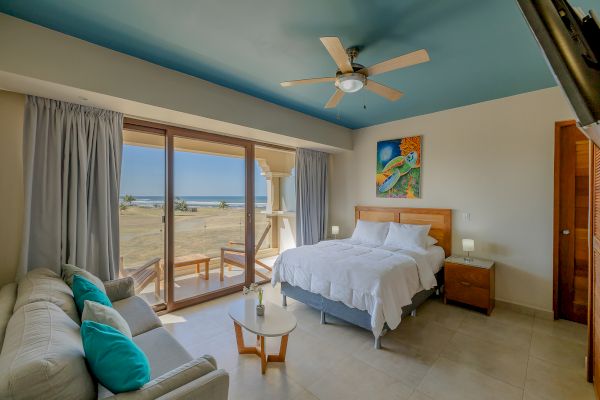 A cozy beachside bedroom with a queen bed, sofa, coffee table, ceiling fan, vibrant painting, and a stunning ocean view from large windows ending the sentence.