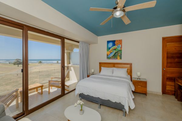 A beachside bedroom with a bed, nightstands, ceiling fan, and artwork on the wall. It has large windows and a door leading to a balcony.