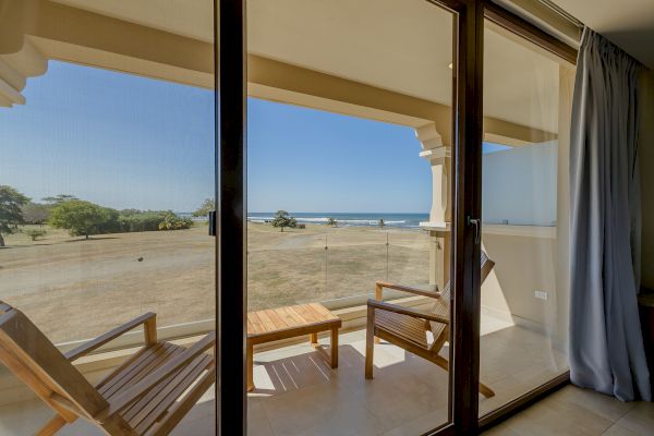The image shows a balcony with two wooden chairs and a table, overlooking a scenic beach with palm trees and clear skies, viewed through glass doors.