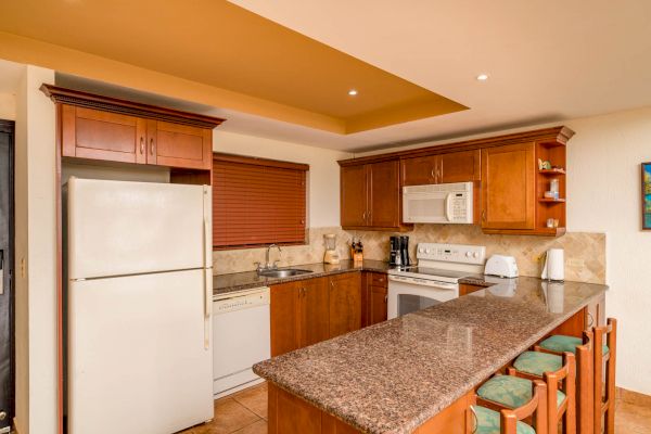 This image shows a kitchen with wooden cabinets, a granite countertop island, and a set of bar stools. It includes a fridge, stove, microwave, and sink.