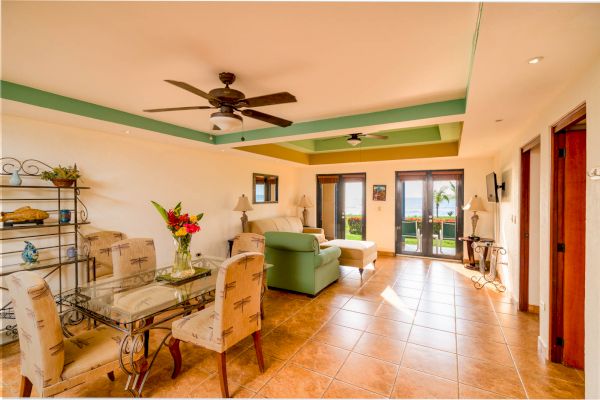 This image shows a bright living and dining area with tiled floors, a ceiling fan, and a glass dining table. There's a view of the ocean through the doors.