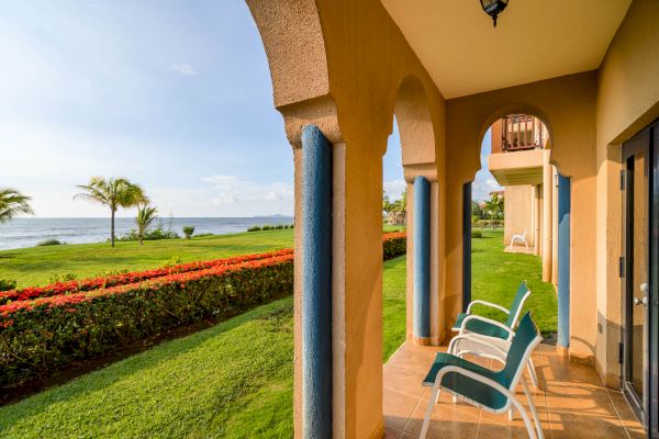 A seaside patio with arches, two chairs, and a serene view of a lush green lawn, palm trees, and the ocean in the distance.