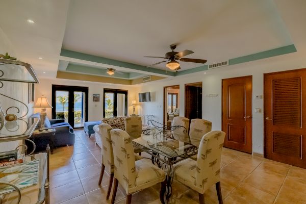 A well-furnished living and dining area with a ceiling fan, glass table, upholstered chairs, and glass doors opening to a balcony overlooking a scenic view.