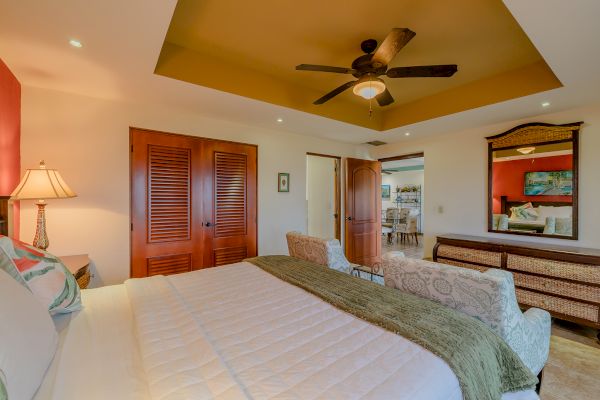 A cozy bedroom with a large bed, ceiling fan, wooden closet doors, mirrored dresser, and an open door leading to another room in the house.