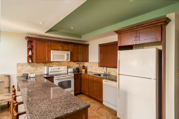 A modern kitchen with wooden cabinets, granite countertop, white appliances including a refrigerator, oven, microwave, and a small dining area.