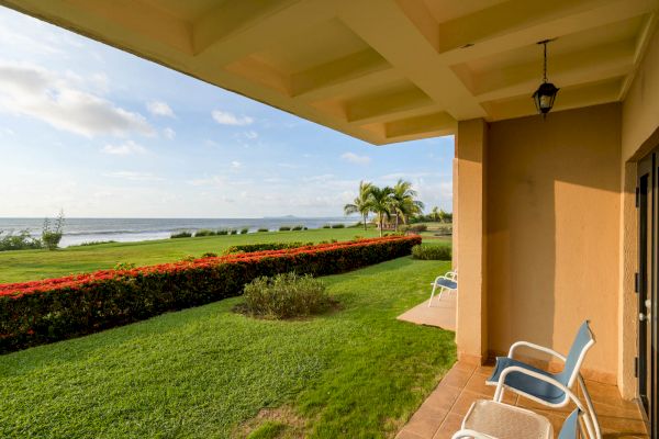 A patio overlooking a lush green lawn, colorful hedges, palm trees, and a distant ocean view, with chairs set for relaxation.