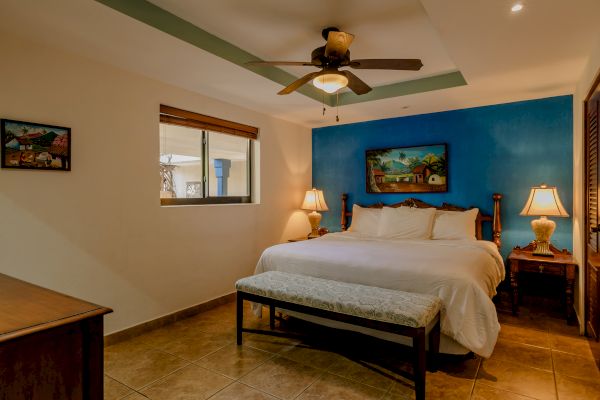A cozy bedroom features a large bed, blue accent wall, ceiling fan, wooden furniture, bedside lamps, bench, and paintings, under warm lighting.