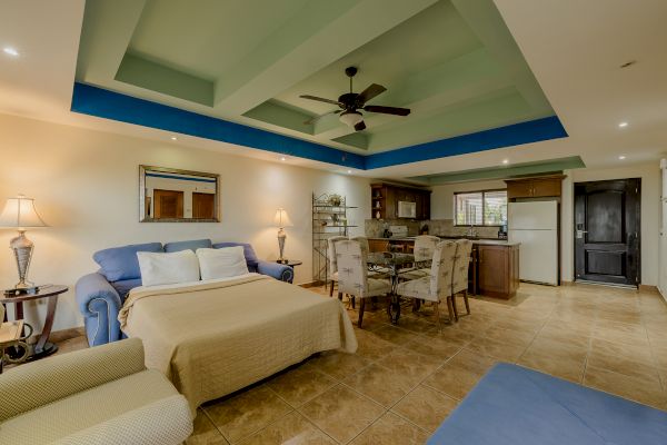 This image shows a spacious living area with a bed, sofa, dining table, ceiling fan, and an open kitchen. The room features a coffered ceiling.