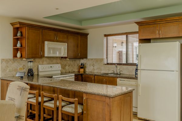 A kitchen features wooden cabinetry, a marble countertop, white appliances including a refrigerator and stove, a sink, and three bar stools.