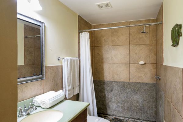 This image shows a bathroom with a sink, mirror, toilet, and a shower area with tiles. Towels are hanging near the sink, and there's a bath mat.