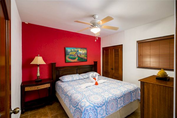 This image shows a cozy bedroom with a red accent wall, double bed, ceiling fan, side table with lamp, dresser, and painting above the bed.