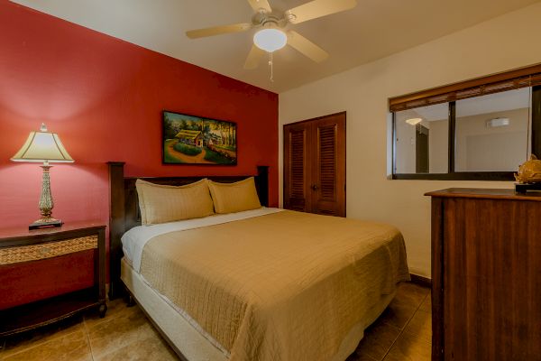 A bedroom with a double bed, two side tables, a ceiling fan, window, painting, and a dresser with a decorative item on it.