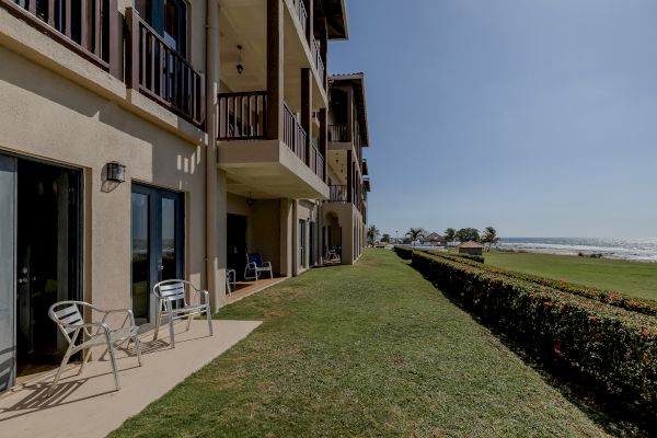 A row of beachfront apartments with balconies, patio chairs, a grassy yard, and a hedge, facing a clear sky and sea view.