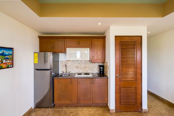 The image shows a small kitchenette with wooden cabinets, a refrigerator, a microwave, a coffee maker, and a stove beside a wooden door.