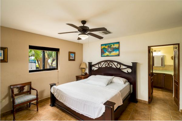 A bedroom with a double bed, wooden headboard, chair, window, ceiling fan, artwork on the wall, and an open door leading to a bathroom.