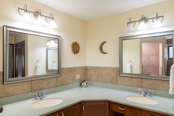 The image shows a bathroom with two sinks on a corner countertop, two mirrors, wall-mounted lights, and wooden cabinets.