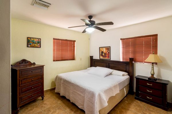 A bedroom with a double bed, wooden furniture, two paintings, and a ceiling fan. It has tiled flooring and windows with pink blinds.